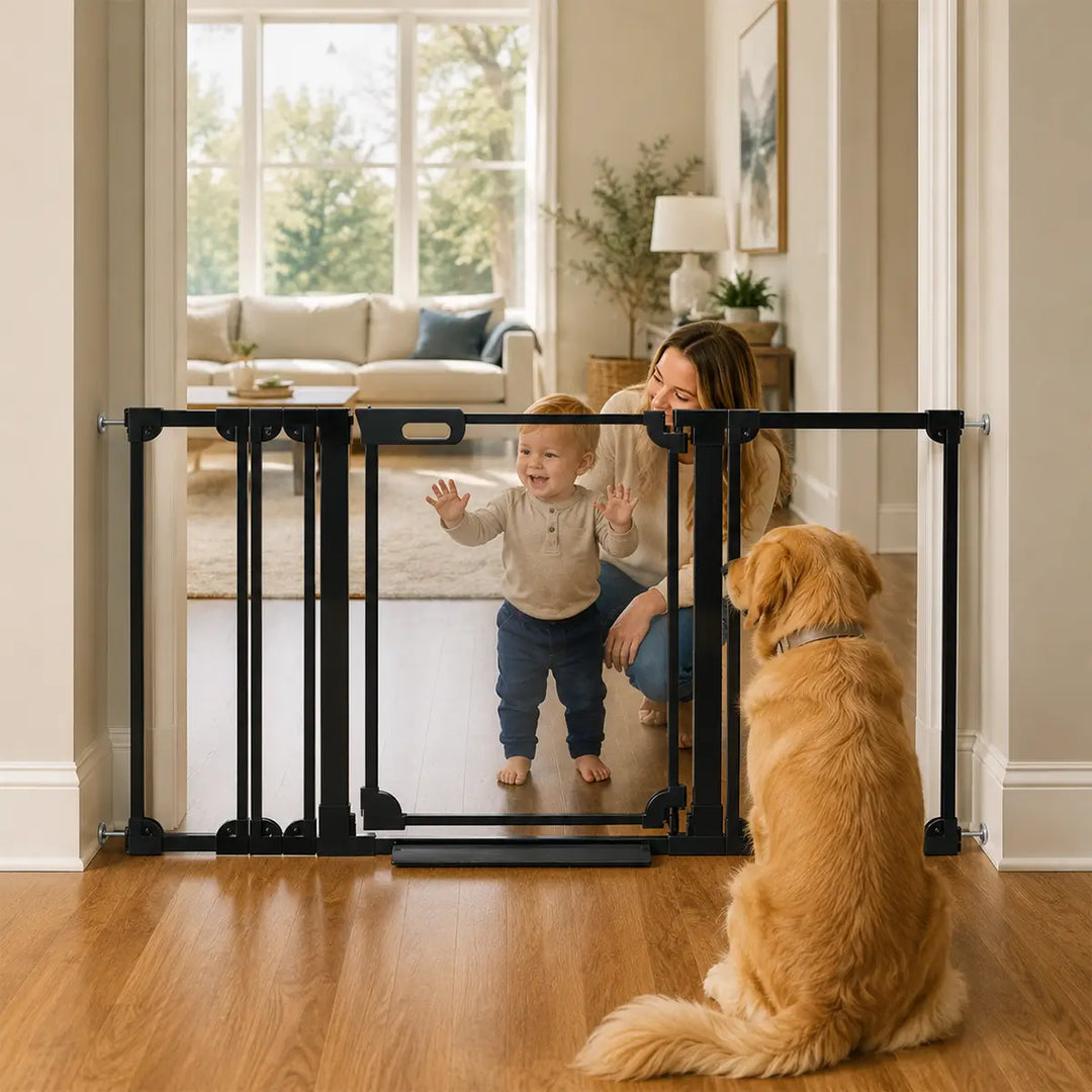 A black metal toddler safety gate installed across a doorway in a bright living room, with a woman crouching behind it and a golden retriever sitting in the foreground.