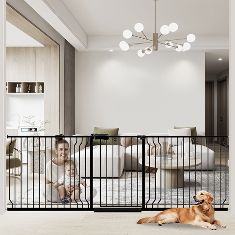 An extra wide baby gate installed in a home, with a black metal frame and a child sitting on a couch in the background. A dog is lying down in front of the gate.