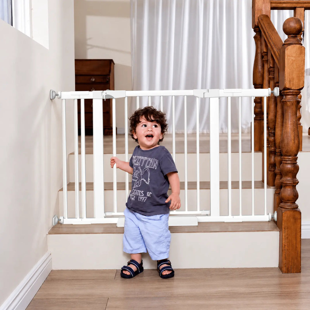 White metal baby safety gate installed at the bottom of stairs with a toddler standing nearby.