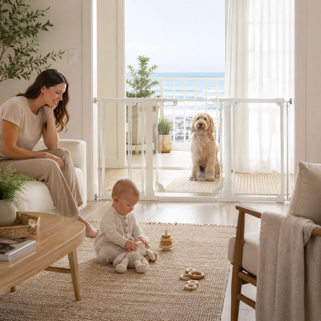 A white baby safety gate installed across a large glass door in a bright living room, with a woman sitting on a chair to the left and a golden dog standing on the balcony beyond the gate.