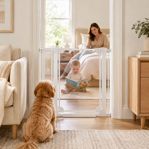 A white adjustable baby gate with a safety latch is installed across a doorway in a bright, modern bedroom. A baby is sitting on the floor reading a book, and a woman is sitting on the bed.